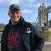 Matthew in coat and green baseball hat poses in front of London Bridge