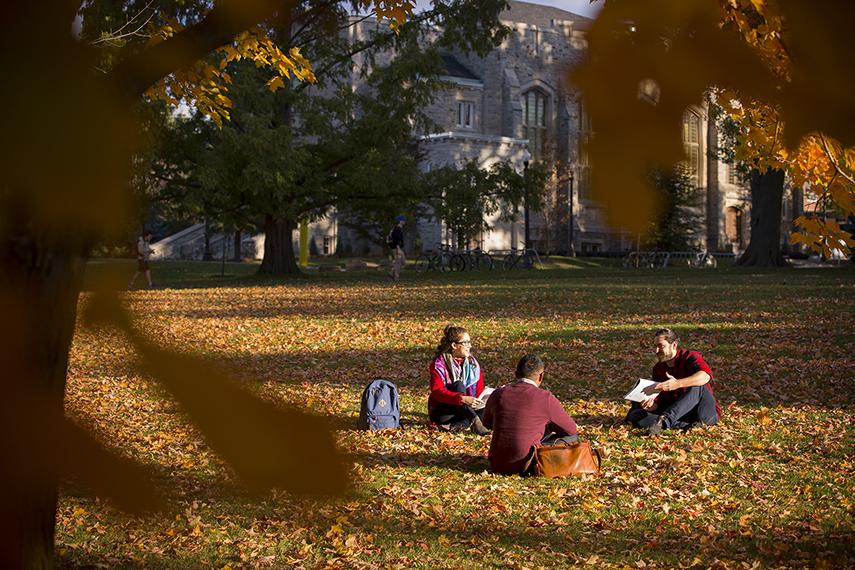 Three students sit and converse on the grass with War Memorial Hall behind them.