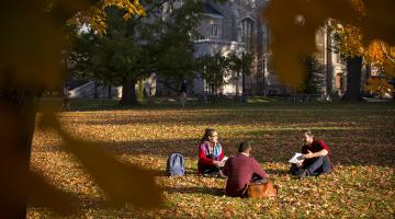 Three students sit and converse on the grass with War Memorial Hall behind them.